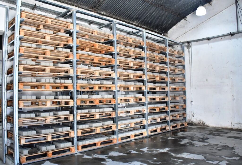 alt="Estanterías metálicas de gran altura con pallets llenos de adoquines grises en la planta municipal de adoquines"
