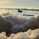 alt="Primer plano de espuma blanca y densa a la orilla del río con un bote difuso en el fondo bajo el sol de la mañana"