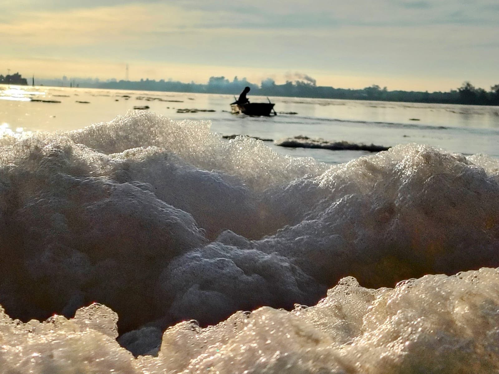 alt="Primer plano de espuma blanca y densa a la orilla del río con un bote difuso en el fondo bajo el sol de la mañana"