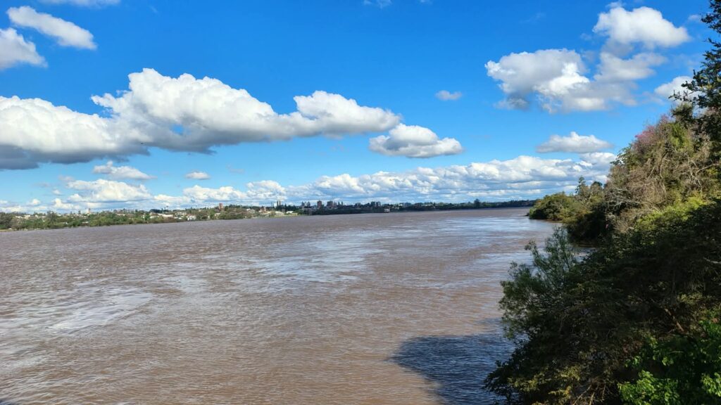 alt="Río Uruguay de aguas marrones con vegetación en la costa y edificios al fondo, en la crecida del río Uruguay en Concordia"